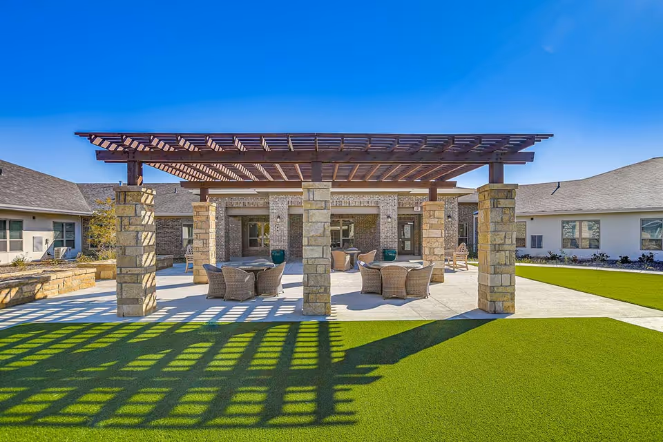 Outdoor patio area at Ariel Pointe Of Sachse featuring a wooden pergola supported by stone pillars, with several round tables and wicker chairs underneath. The patio is surrounded by green artificial turf and buildings with windows under a clear blue sky.