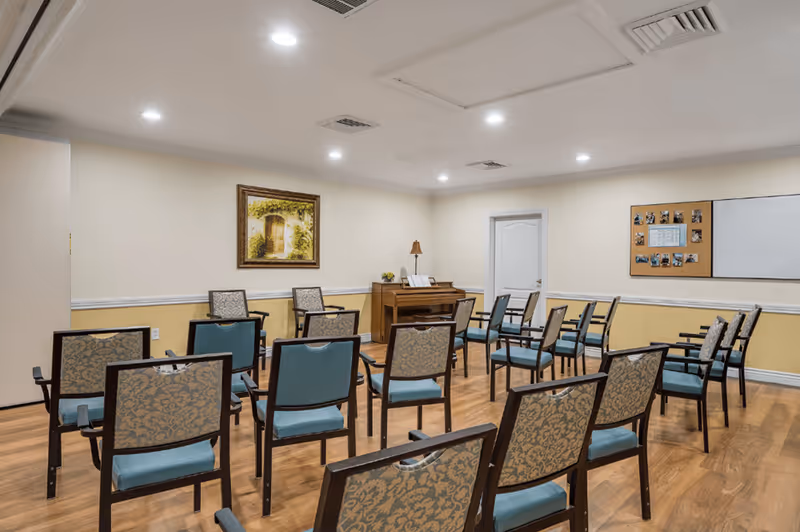 A small meeting or activity room with several chairs arranged in rows facing a piano against the wall. The room has light-colored walls with a framed picture and a bulletin board. The floor is wooden, and the ceiling has recessed lighting.