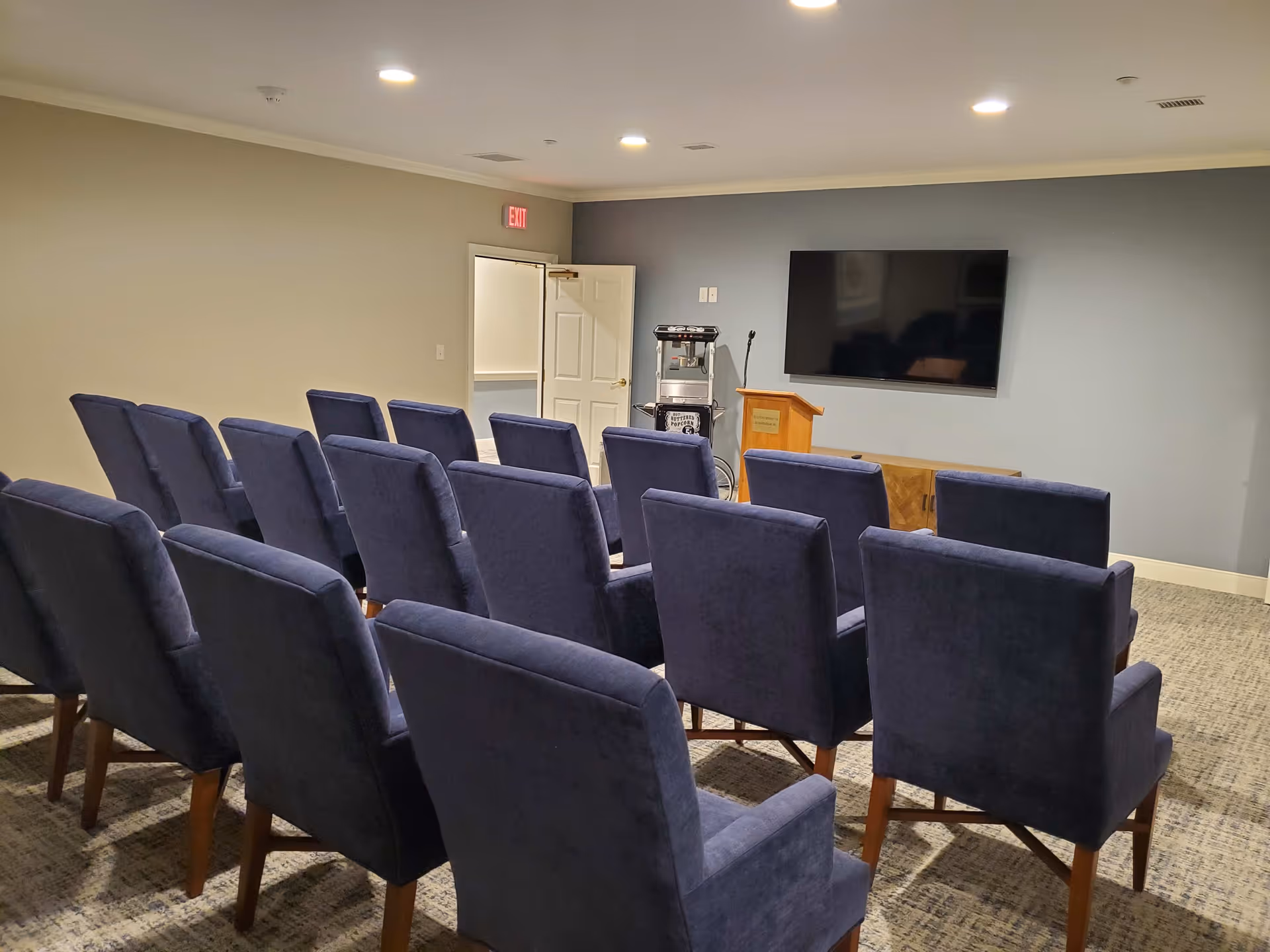 A small conference or presentation room with three rows of blue upholstered chairs facing a wall-mounted flat screen TV. There is a wooden podium and a popcorn machine near the TV. The room has beige and gray walls, carpeted floor, and recessed ceiling lights.