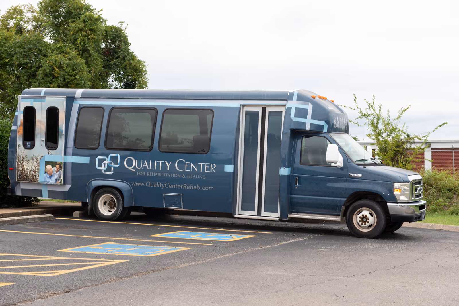 A blue shuttle bus parked in a parking lot with two handicap parking spaces visible in front. The bus has the logo and text 'Quality Center for Rehabilitation & Healing' along with the website 'www.QualityCenterRehab.com' on its side. There is a small image of two elderly people on the rear side of the bus. Trees and a building are visible in the background.