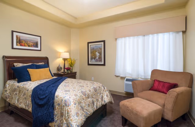 A cozy bedroom in a senior living facility with a single bed covered in a patterned bedspread, blue and yellow pillows, and a blue throw blanket. Next to the bed is a wooden nightstand with a lamp, a vase with yellow flowers, and a small decorative item. There are two framed pictures on the light yellow walls. A large window with white curtains is beside a comfortable brown armchair with a matching ottoman and a red pillow.