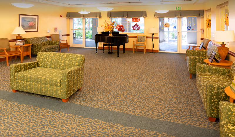 A spacious and well-lit common area with patterned green armchairs and sofas arranged around the room. There is a black grand piano decorated with a floral arrangement in the center near the windows. The windows and glass doors have seasonal fall decorations including pumpkins and leaves. The room has beige walls, patterned carpet, and several wooden side tables with lamps and framed pictures.