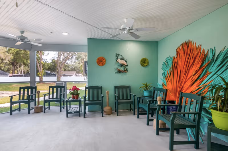 Covered outdoor seating area with green wooden chairs, potted plants, ceiling fans, and a colorful wall mural.