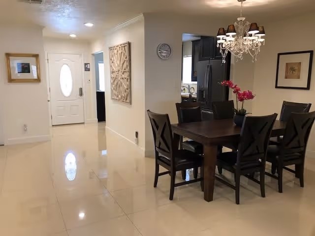 Interior view of a dining area with a dark wooden table and six matching chairs. A chandelier with multiple lights hangs above the table, and a pink orchid centerpiece is on the table. The room has light-colored tiled flooring and cream-colored walls. To the left, there is a hallway leading to a white front door with an oval window. Wall decorations include a framed picture and a decorative wall panel.