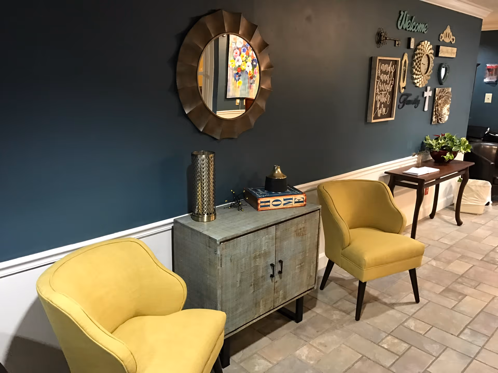 A seating area with two yellow armchairs, a small wooden cabinet, decorative mirror, and wall art on a dark teal wall.