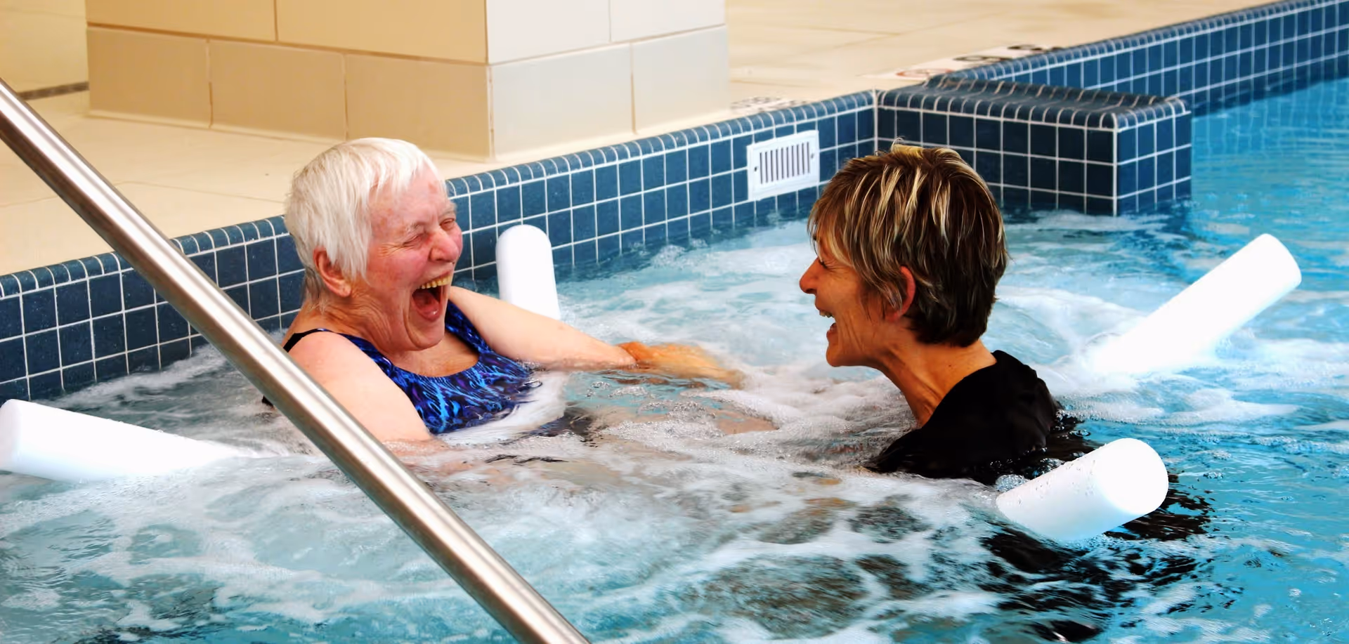 Two women enjoying a bubbly indoor pool or hot tub, laughing and facing each other. One woman has short white hair and is wearing a blue swimsuit, while the other has short brown hair and is wearing a black top. The pool has blue tiled edges and white pool noodles floating in the water.