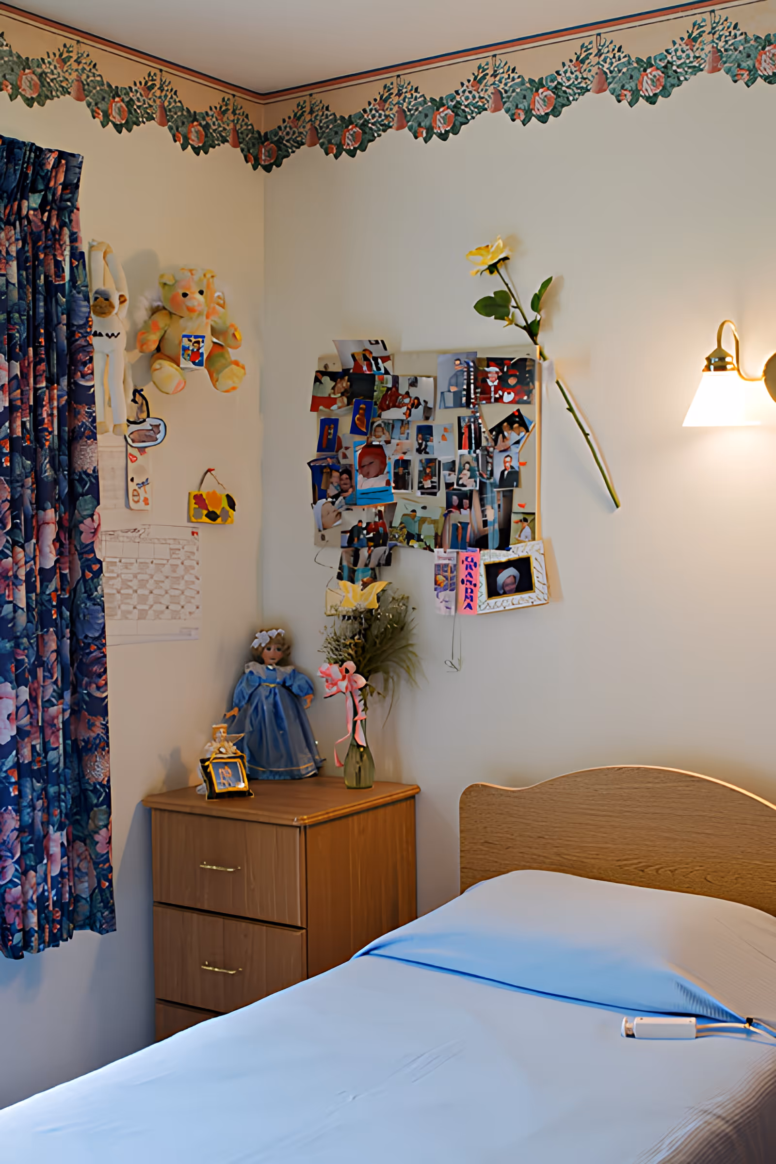 A cozy bedroom corner in the Ansted Center featuring a single bed with a light blue sheet and a wooden headboard. Next to the bed is a wooden nightstand with two drawers, decorated with a doll in a blue dress, a small framed photo, and a vase with flowers. The wall above the nightstand displays a collage of personal photos and a single yellow flower in a vase. The room has floral patterned curtains and a floral border near the ceiling. A wall-mounted lamp provides soft lighting.