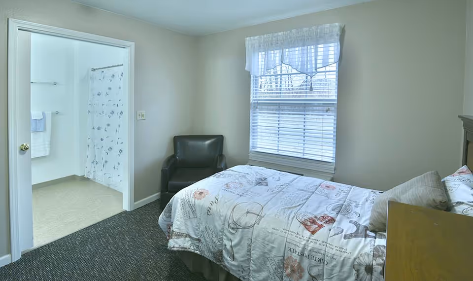 A small bedroom with a single bed covered in a patterned comforter, a black armchair in the corner, and a window with white blinds and a sheer valance. An open door reveals a bathroom with a shower curtain, towel rack, and tiled floor.
