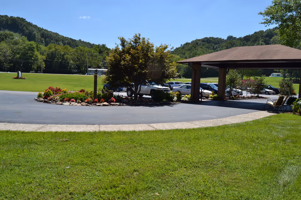 Covered porte-cochere entrance with a circular driveway, flowerbeds, parked cars, and surrounding grassy hills.