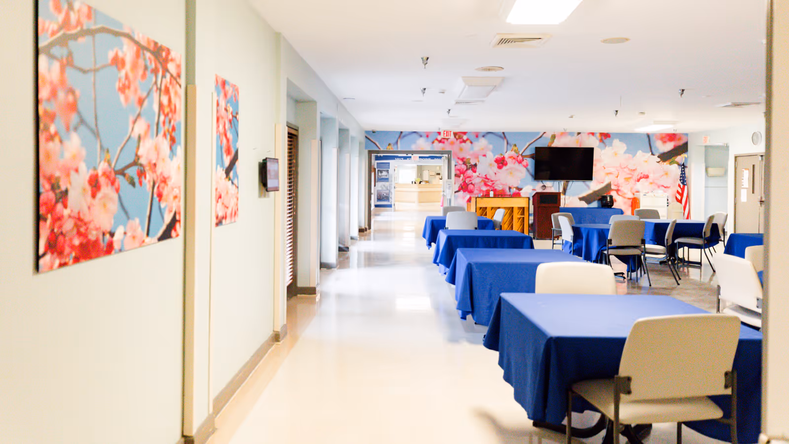 Interior view of a senior living facility hallway leading to a common area with tables covered in blue tablecloths and chairs arranged around them. The walls are decorated with large floral artwork featuring pink blossoms on a blue background. A television and a piano are visible in the common area, along with an American flag in the corner.