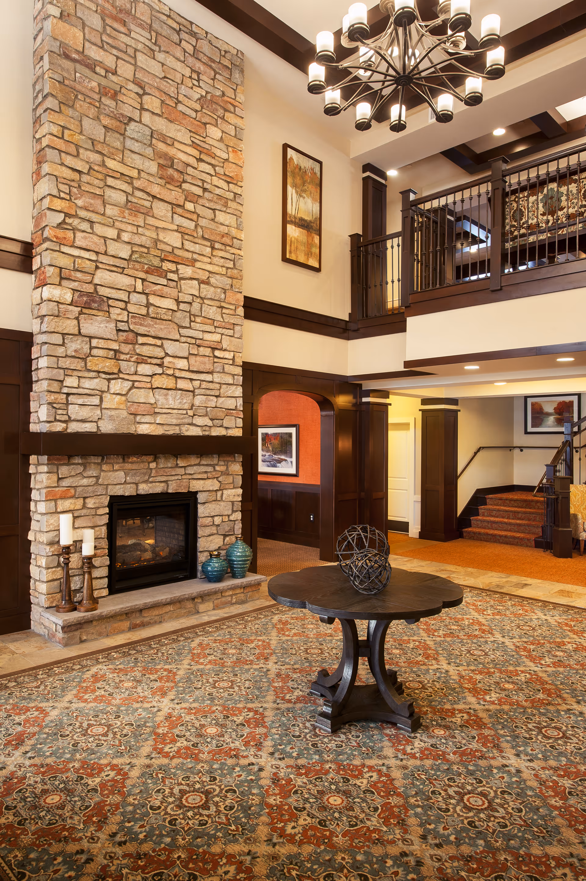 A spacious senior living facility common area featuring a tall stone fireplace with candles and decorative vases on the hearth. The room has a large patterned carpet, a round wooden table with a decorative metal sphere centerpiece, and a chandelier hanging from a high ceiling with exposed wooden beams. There is a staircase leading to an upper level with a railing, and framed artwork on the walls.