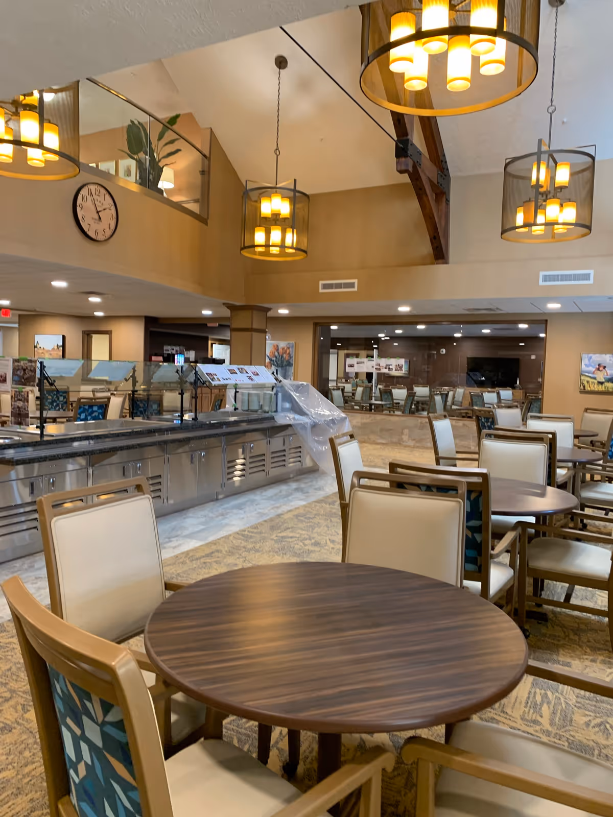 Interior view of a dining area in a retirement community with round wooden tables and cushioned chairs. The room features warm lighting with hanging pendant lamps, a high ceiling with exposed wooden beams, and a clock on the wall. There is a serving counter with a protective glass barrier and some covered items, and additional seating is visible in the background.