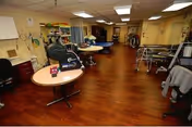 Interior view of a healthcare facility room with wooden flooring, several round tables, chairs, medical equipment, and shelves with supplies. The room is well-lit with ceiling lights and has a clinical, organized appearance.