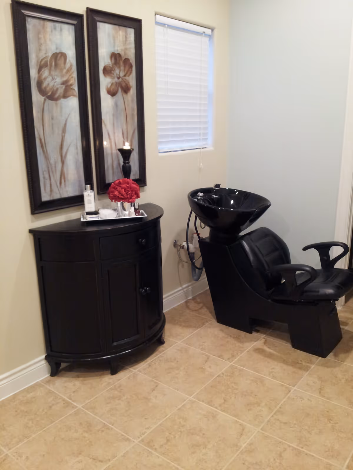 A small hair washing station with a black salon chair and black sink basin. Next to it is a black curved cabinet with a tray holding a bottle, a candle, and a red flower arrangement. Two framed floral paintings hang on the wall above the cabinet, and a small window with closed blinds is visible.