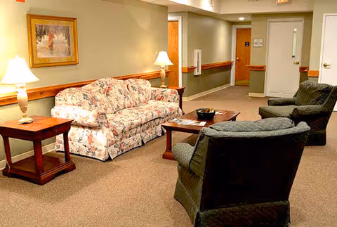 Carpeted lounge area with a floral sofa, two upholstered armchairs, wooden side and coffee tables, lamps, and a hallway entrance.