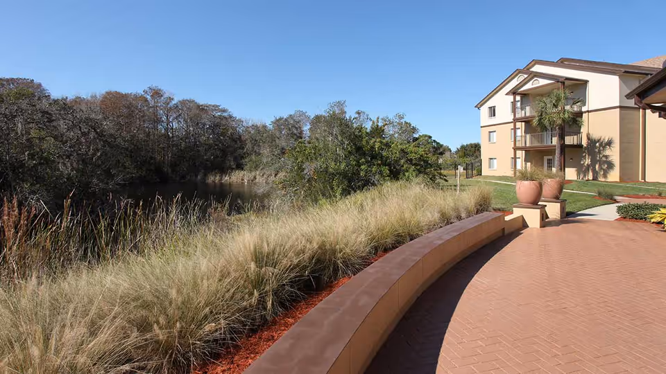 Curved brick patio and landscaped grasses beside a pond with a multi-story beige residential building in the background under a clear blue sky.