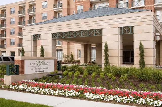 Exterior view of The Village of Tanglewood building entrance with a covered drop-off area, surrounded by landscaped flower beds with red, white, and pink flowers and green shrubs.