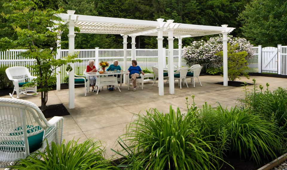 Three elderly people sitting and chatting at a white outdoor table under a white pergola in a garden area with green plants, white fencing, and blooming bushes.