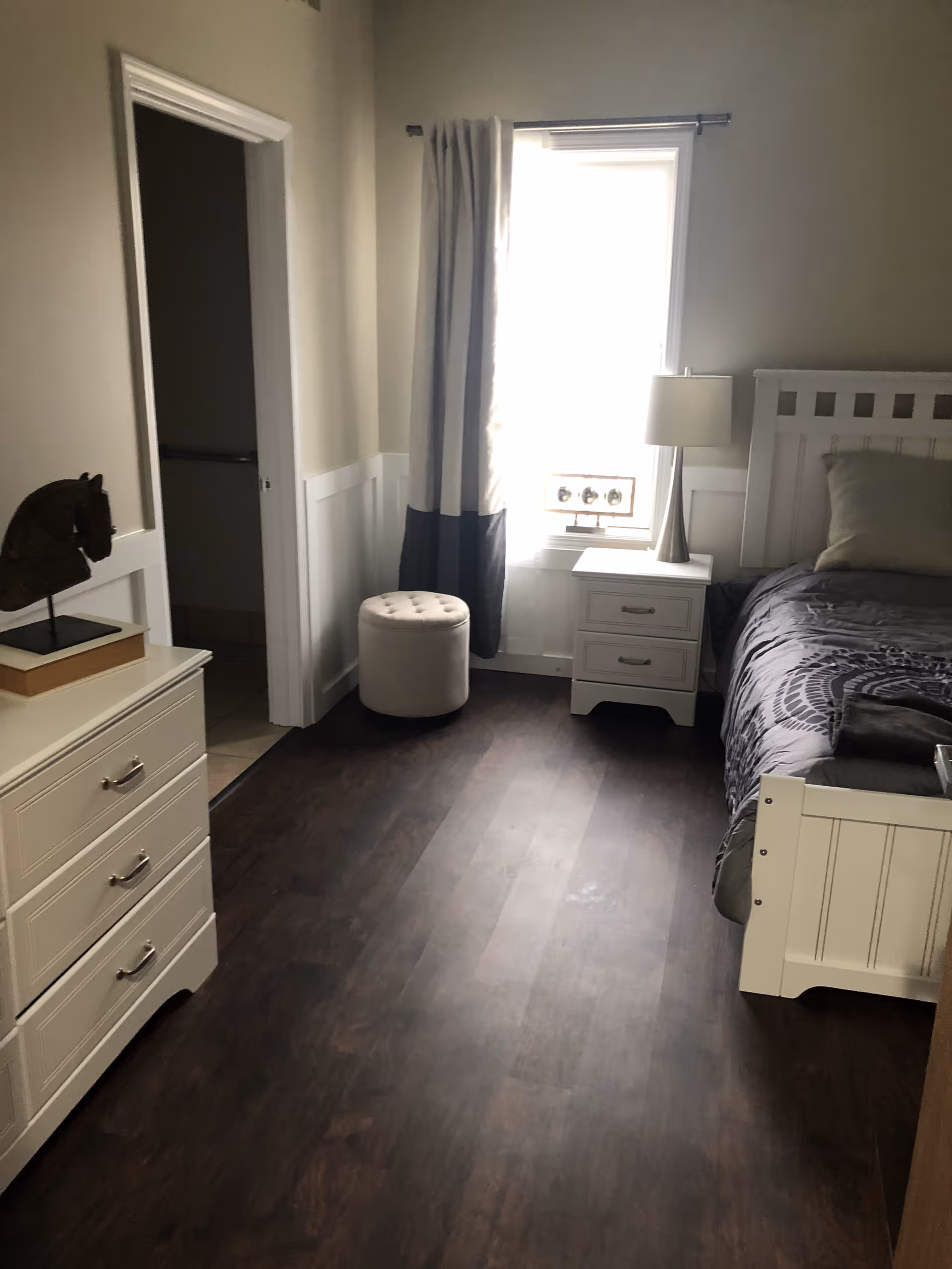 A bedroom with dark wood flooring, a white bed with gray bedding, a white nightstand with a lamp, a window with curtains, a white dresser with a decorative horse head sculpture, and an open doorway leading to a bathroom.