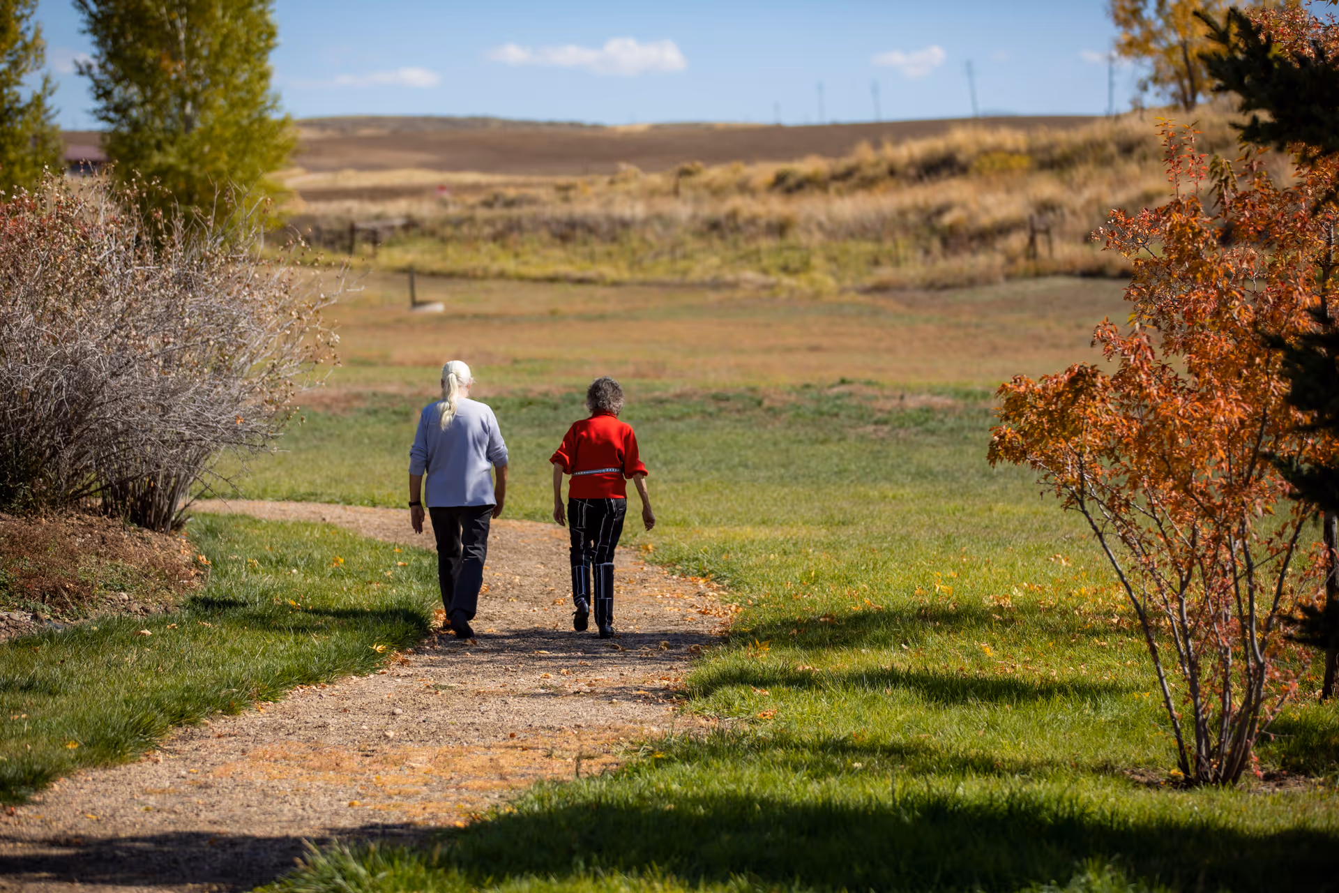 Two elderly women walking on a gravel path through a grassy area with trees showing autumn colors, under a clear blue sky.