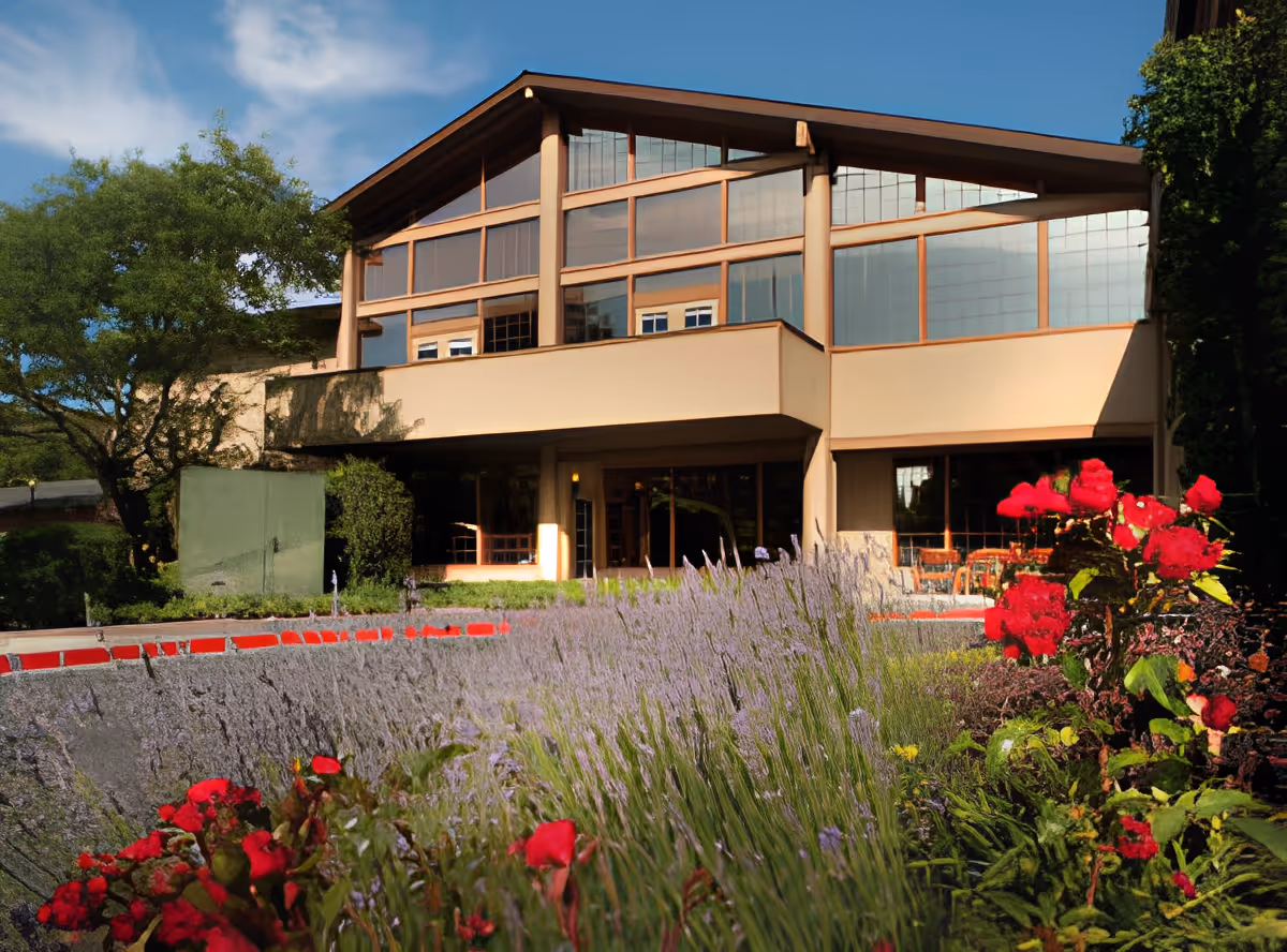 Exterior view of a modern building with large windows and a slanted roof, surrounded by lush greenery and vibrant red flowers in the foreground under a blue sky.