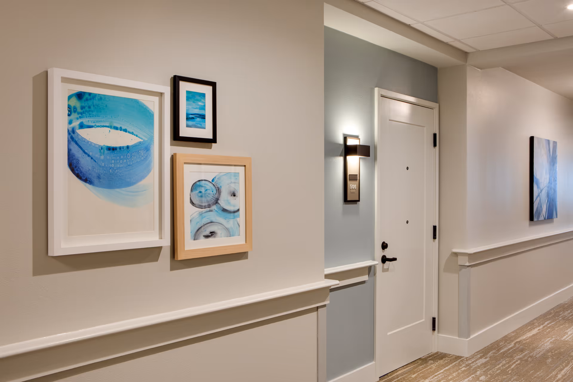 Well-lit interior hallway with framed blue artwork and a white apartment door with a wall sconce.