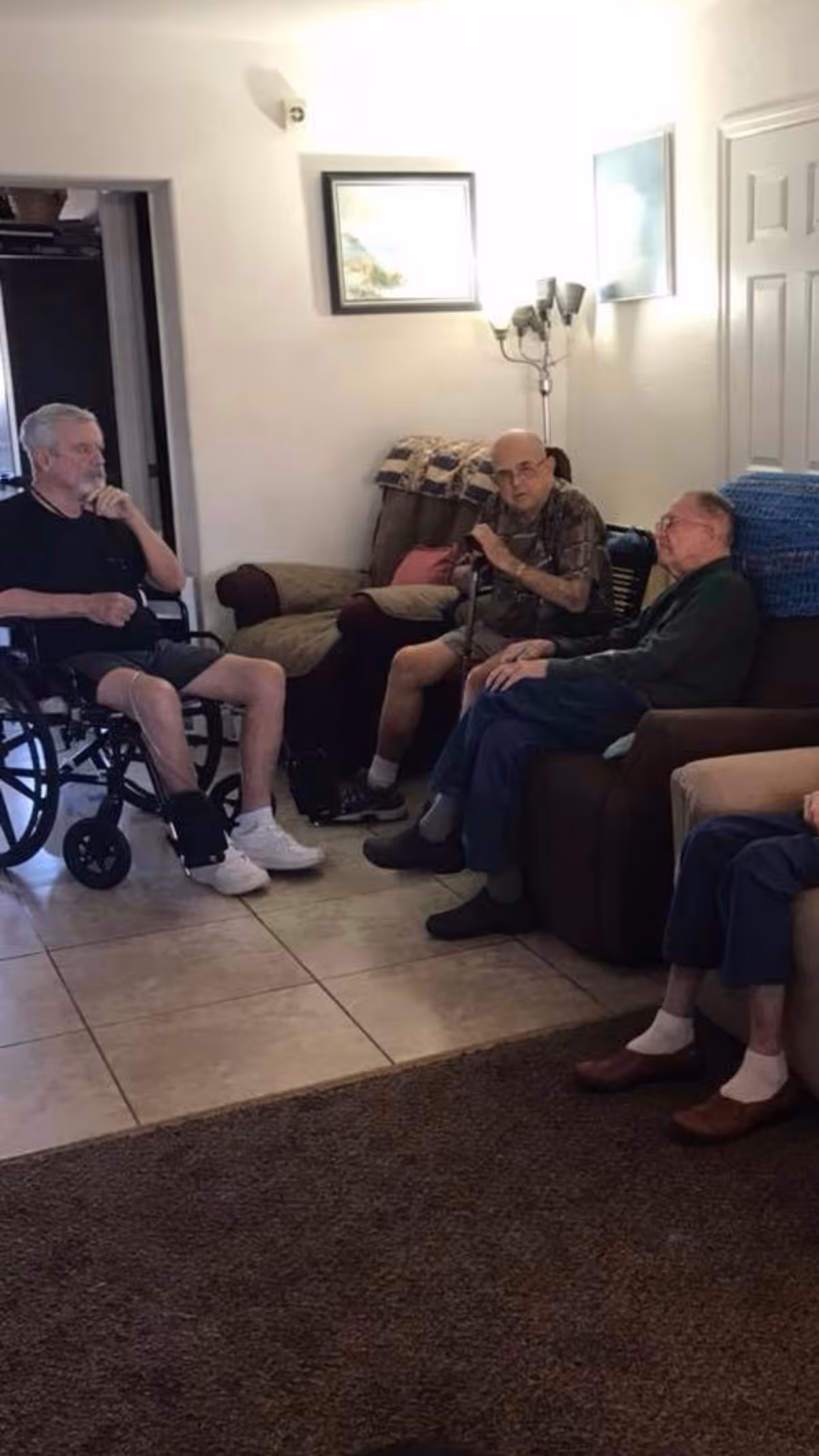 A group of elderly men sitting and conversing in a living room area. One man is in a wheelchair, another is holding a cane, and two others are seated on armchairs. The room has tiled flooring, a carpet, framed pictures on the wall, and a standing lamp.
