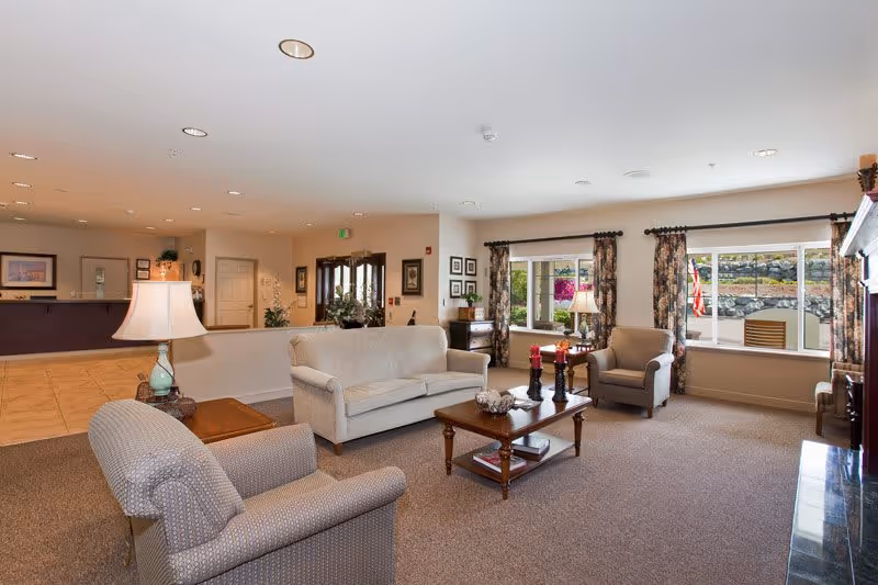 A bright and spacious senior living facility common area with beige carpeting and neutral-colored walls. The room features a beige sofa, two armchairs, a wooden coffee table with decorative items, and a side table with a lamp. Large windows with floral curtains allow natural light to fill the space. In the background, there is a reception desk and a hallway leading to other parts of the facility.