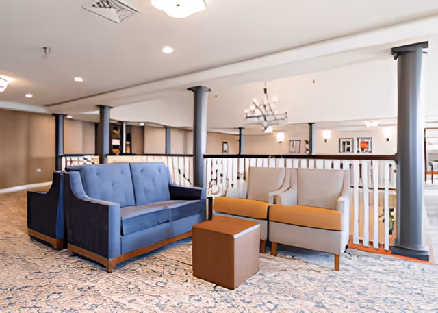 A seating area in a senior living facility with a blue sofa, two beige armchairs with orange cushions, and a small brown ottoman on a patterned carpet. The space is open with white railings and gray columns, and there are chandeliers and wall sconces providing lighting.