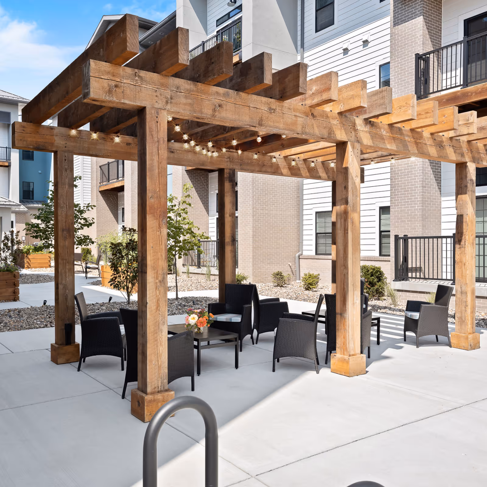 Wooden pergola with string lights covering an outdoor seating area with wicker chairs and tables in a senior living courtyard.