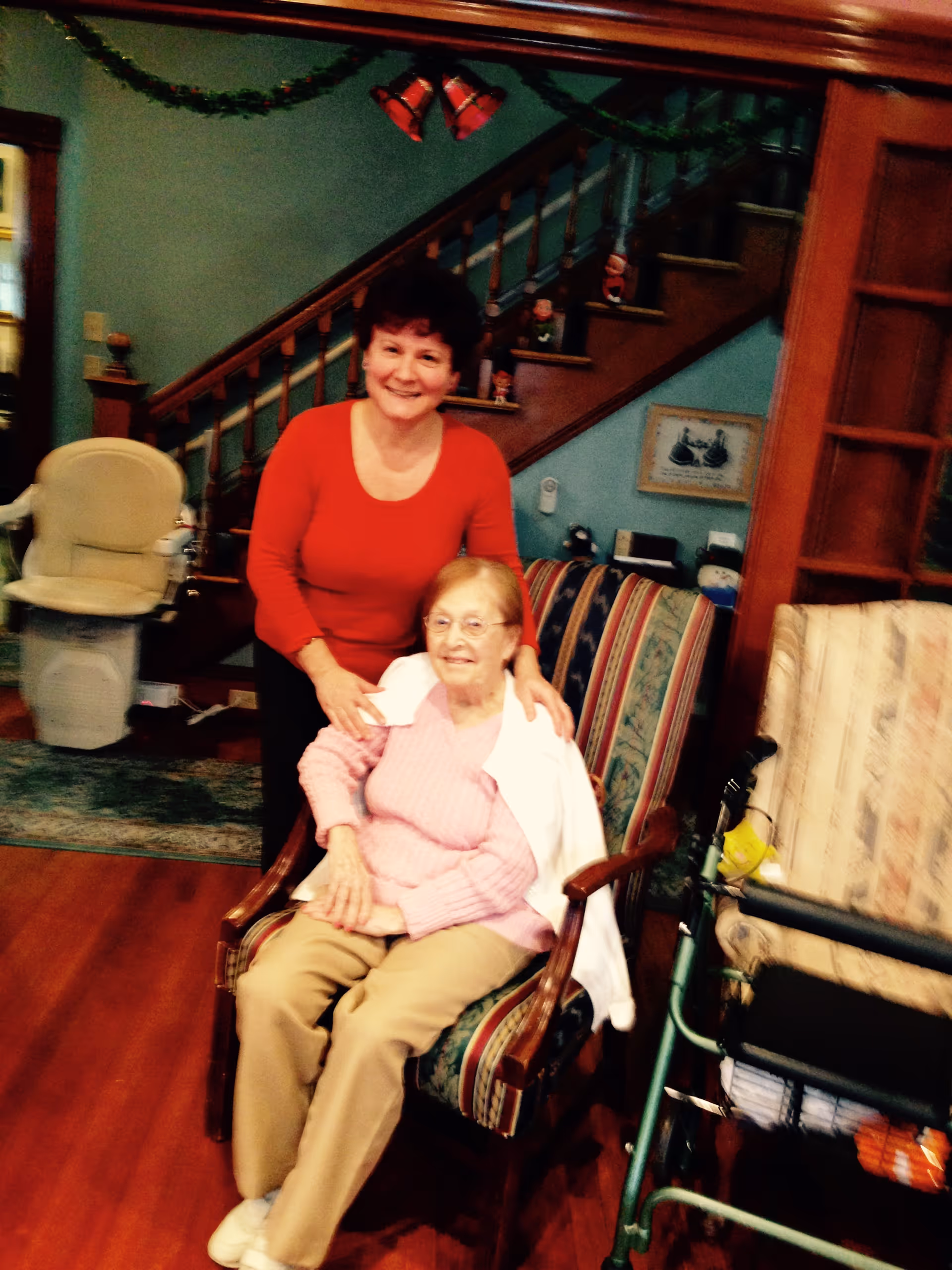 An elderly woman sitting in a striped armchair with a walker nearby, smiling, while a middle-aged woman in a red top stands behind her with hands on her shoulders. They are in a cozy living room with wooden floors, a staircase decorated with garland and bells, and various home decorations.
