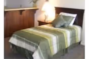 A neatly made single bed with a green and white striped bedspread and two pillows, positioned next to a wooden nightstand with a lamp on it. The room has a carpeted floor and a partial view of a wooden counter or desk in the background.