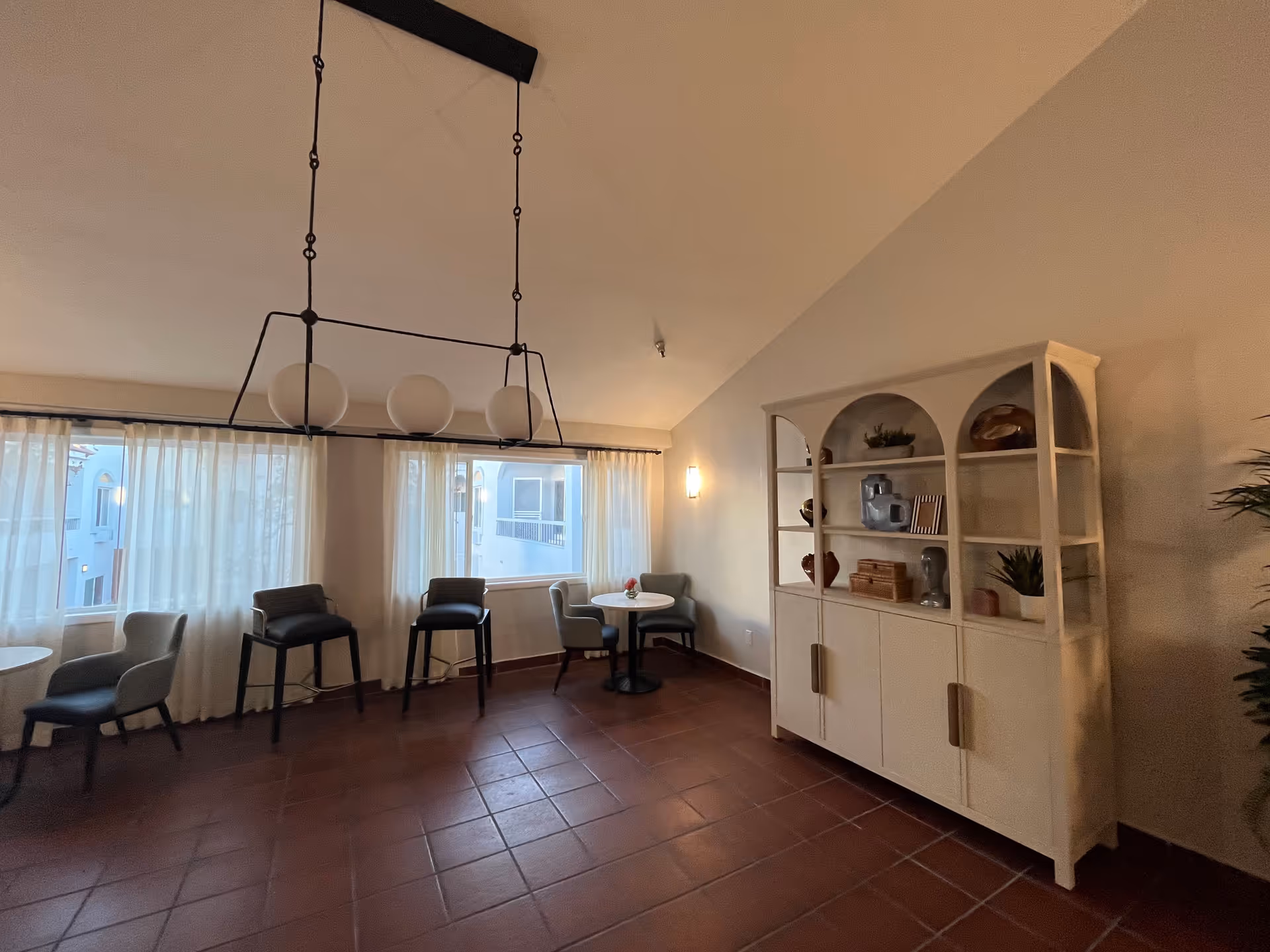 A cozy interior room with terracotta tiled floor, a white cabinet with decorative items on the right wall, and three small round tables each accompanied by two chairs. Large windows with sheer white curtains allow natural light to fill the space. A modern black chandelier with three round white bulbs hangs from the ceiling.
