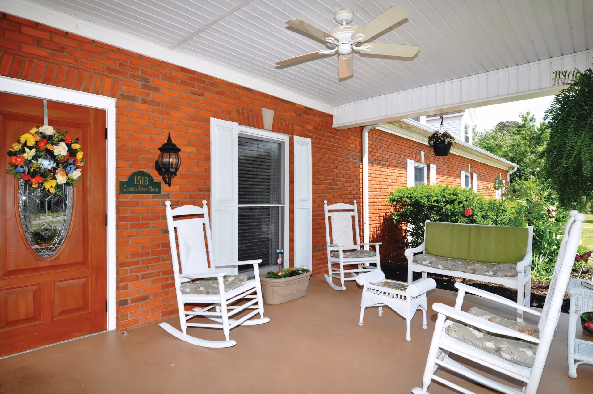 Covered porch area with white rocking chairs, a white bench with green cushion, a small white wicker table, and a hanging plant. The porch has a brick wall with a window and a wooden door decorated with a colorful flower wreath. There is a ceiling fan above and greenery visible outside.