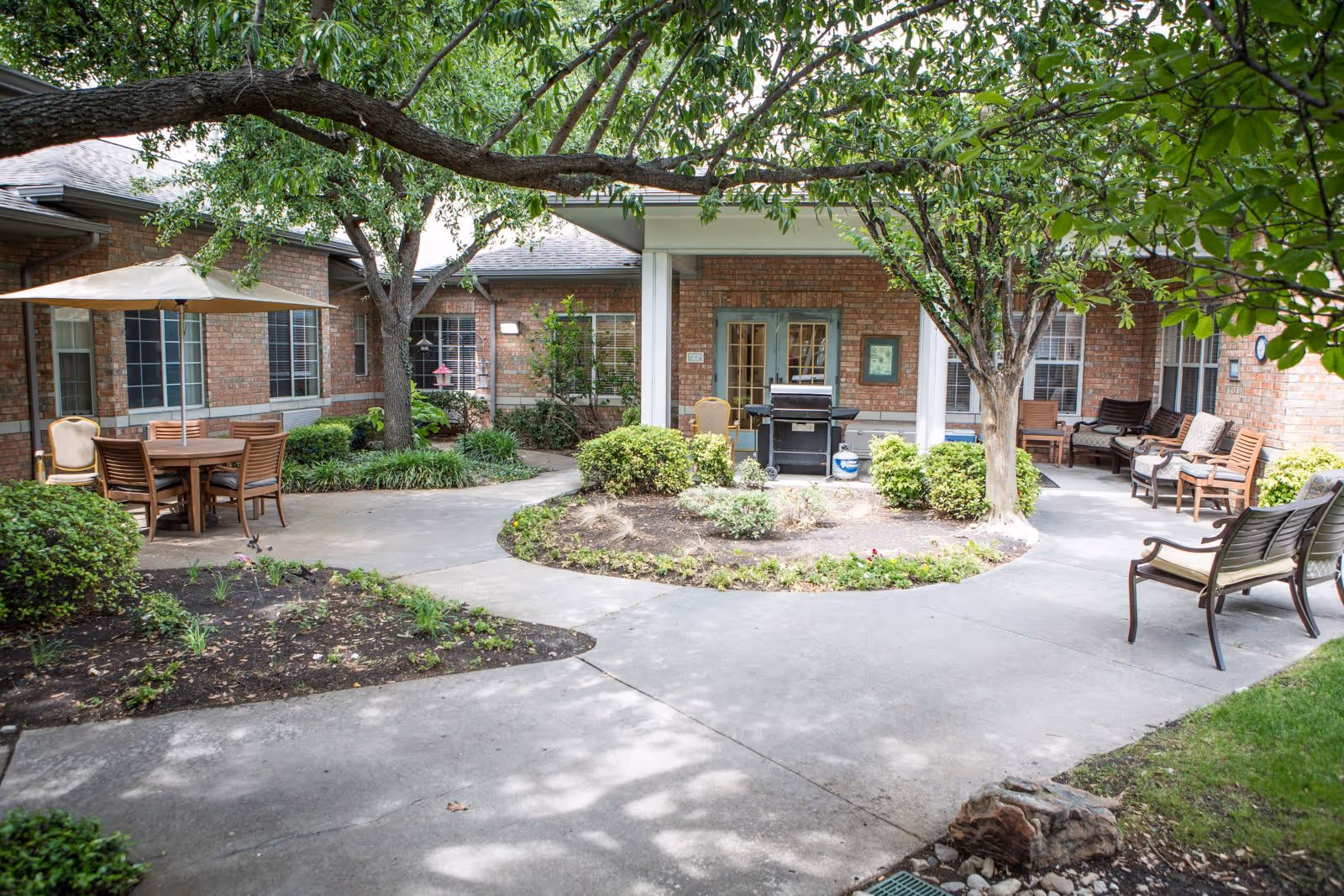 Outdoor courtyard area at Spring Creek Assisted Living featuring a circular concrete walkway surrounding a garden bed with shrubs and small plants. There are several trees providing shade, wooden chairs and benches arranged around the courtyard, and a table with an umbrella and chairs on the left side. The building has brick walls and multiple windows and doors opening into the courtyard.