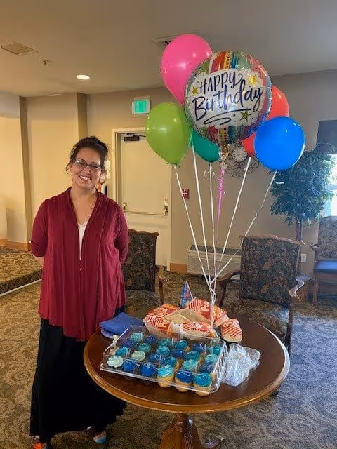 A woman wearing glasses and a red cardigan stands smiling next to a round wooden table with a birthday cake, cupcakes, and colorful balloons including one that says 'Happy Birthday' in a cozy room with patterned carpet and upholstered chairs.