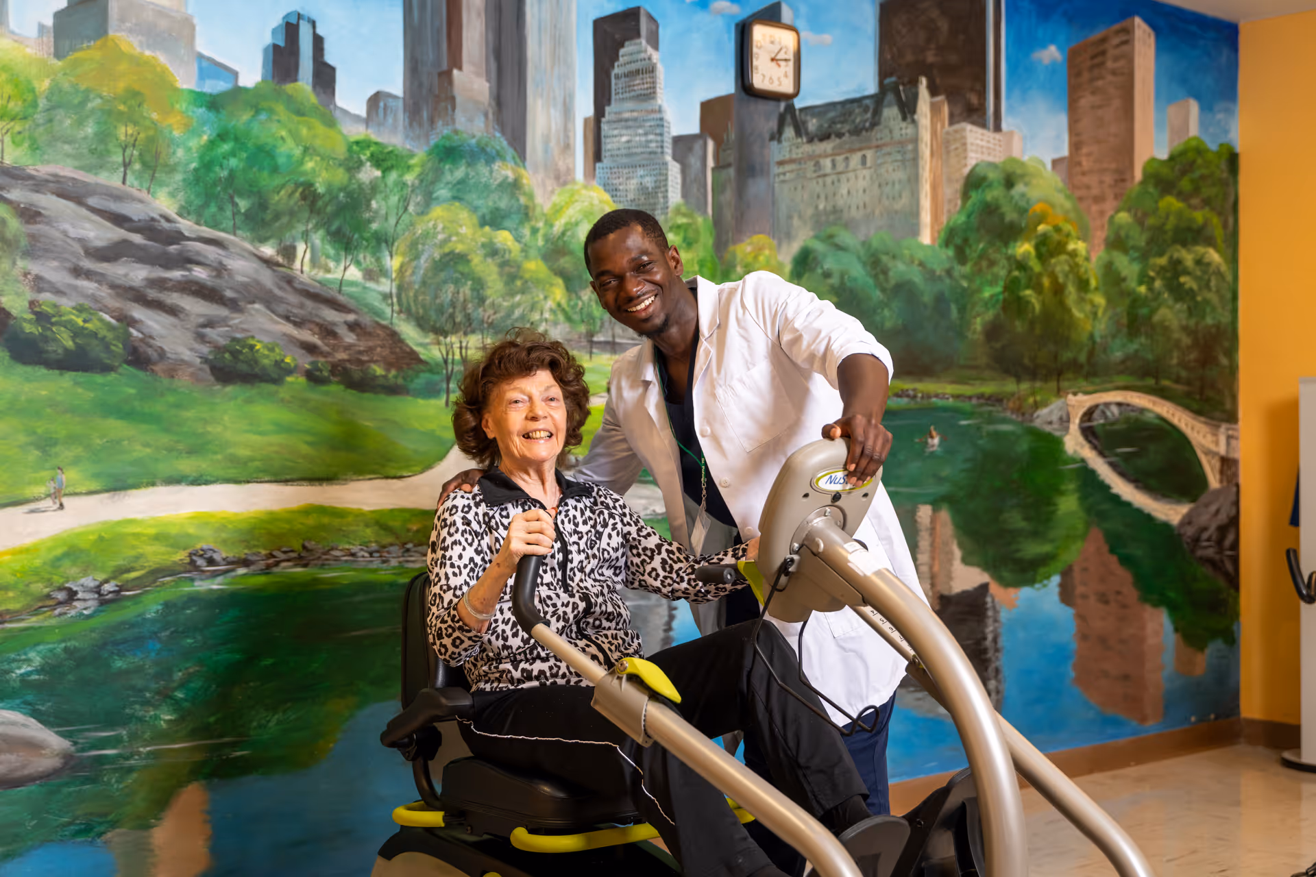 An elderly woman using a seated exercise machine with the assistance of a smiling male healthcare worker in a white coat, in front of a large mural depicting a park scene with trees, a pond, a bridge, and city buildings in the background.