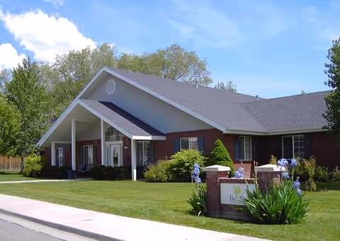 Exterior view of a single-story brick building with a gabled roof, surrounded by green grass, bushes, and trees under a blue sky with some clouds. A sign in front reads 'Bel Aire Assisted Living'.