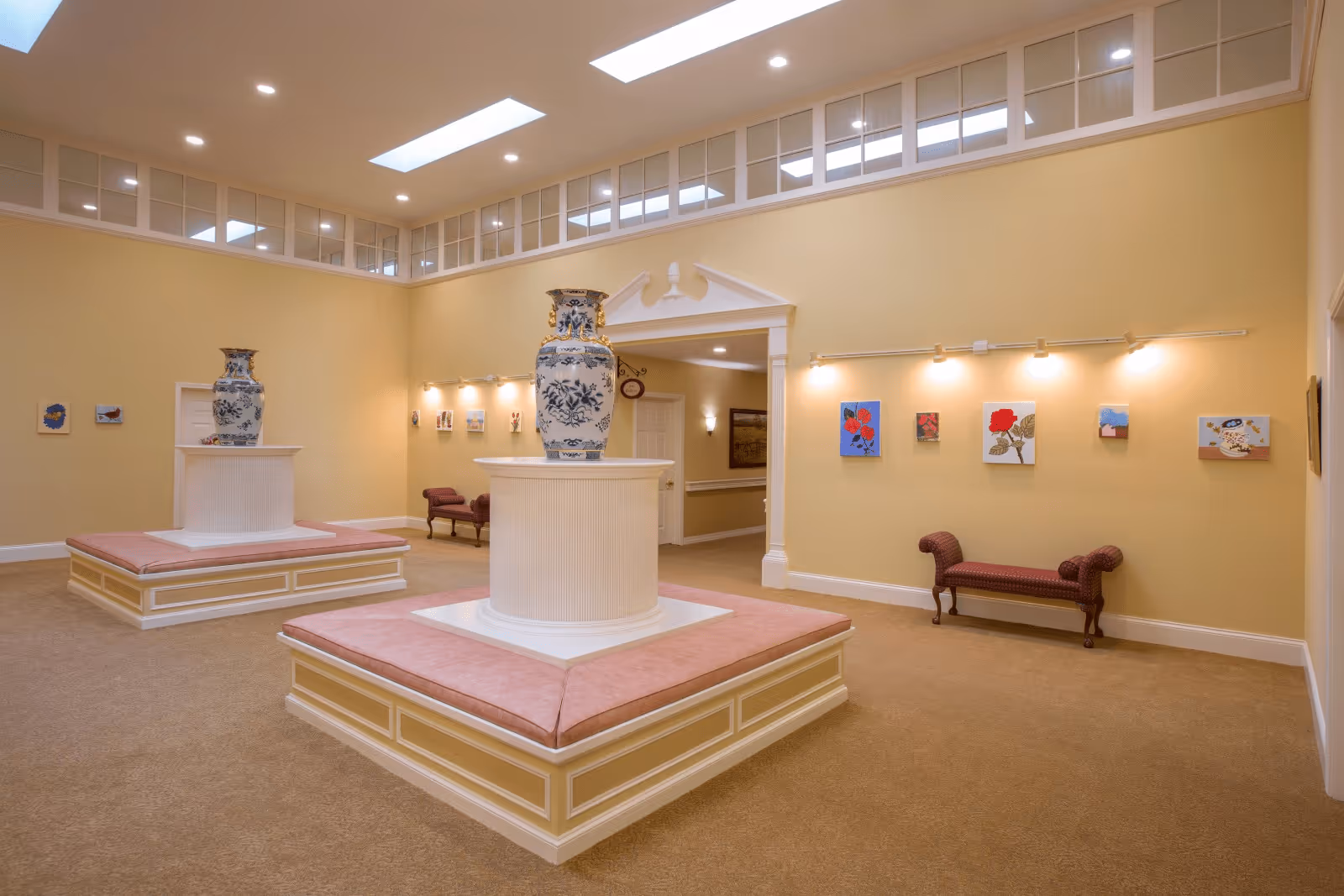 A spacious, well-lit interior room with beige walls and carpeted floor featuring two large decorative vases on white pedestals with pink cushioned seating around them. The walls display small framed artworks illuminated by wall-mounted lights. There are two upholstered benches along the walls and a doorway leading to another area.