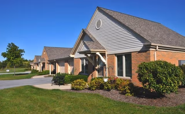 Exterior view of a single-story senior living community building with brick and siding facade, multiple entrances, well-maintained landscaping with bushes and flowers, and a clear blue sky.