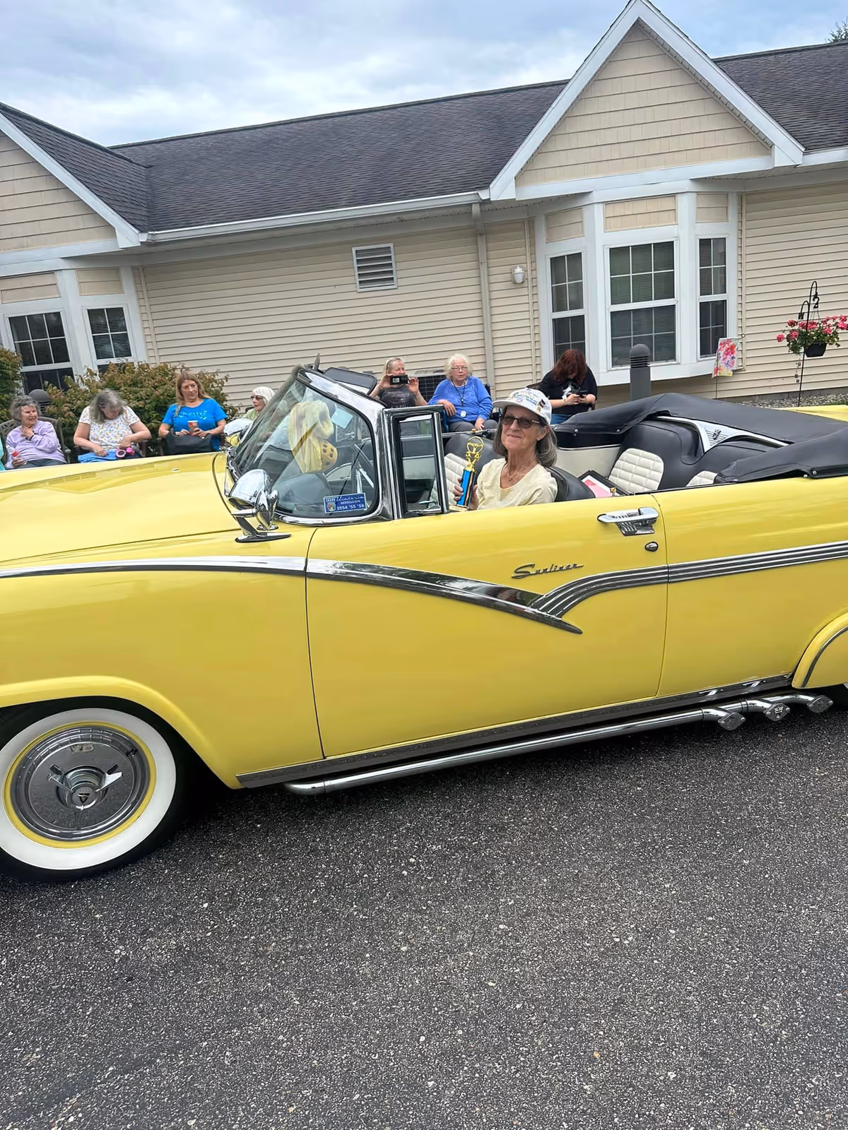 A yellow vintage convertible parked outside a retirement home with a woman holding a trophy inside and several residents seated on benches behind.