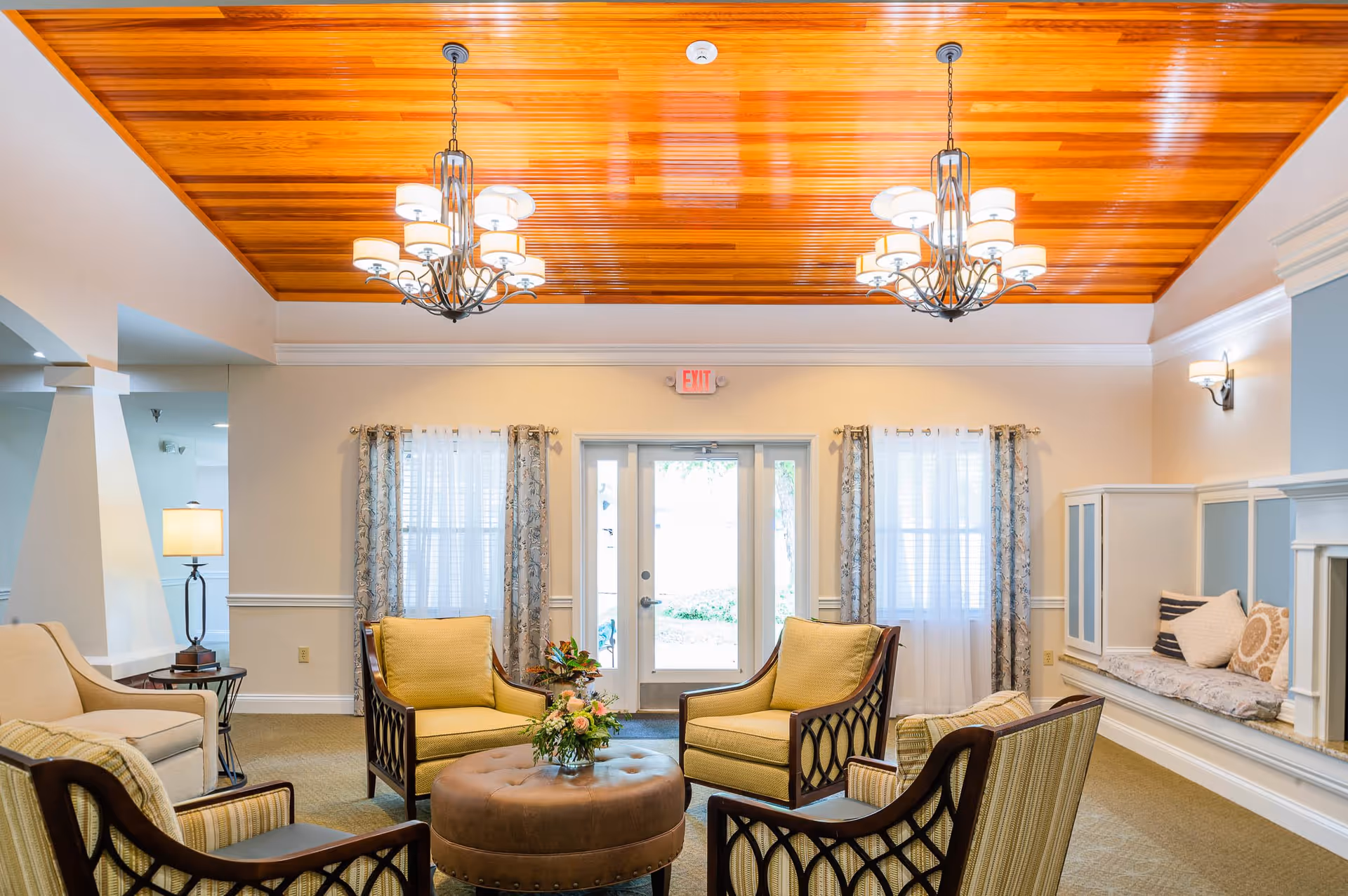 A cozy living room area with four cushioned armchairs arranged around a round leather ottoman with a flower arrangement on top. The room features a polished wooden ceiling with two chandeliers, light beige walls, two windows with sheer curtains, and a glass door leading outside. There is a built-in bench with cushions along the right wall and a table lamp on the left side.