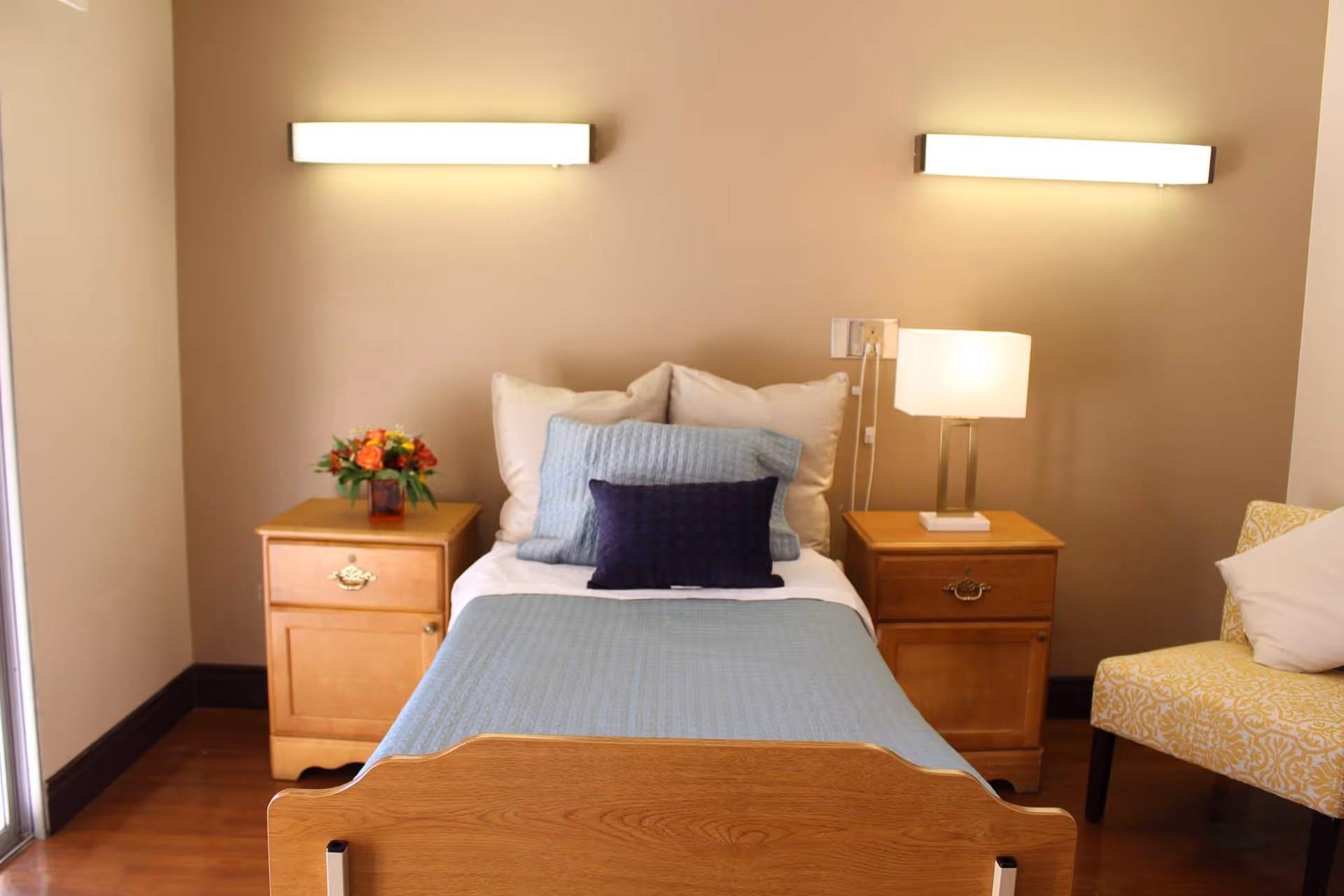 A neatly made single bed with light blue bedding and multiple pillows, flanked by two wooden nightstands. One nightstand has a small bouquet of flowers, and the other has a modern table lamp. A yellow patterned armchair with a white pillow is visible on the right side. The room has beige walls and two rectangular wall lights above the bed.