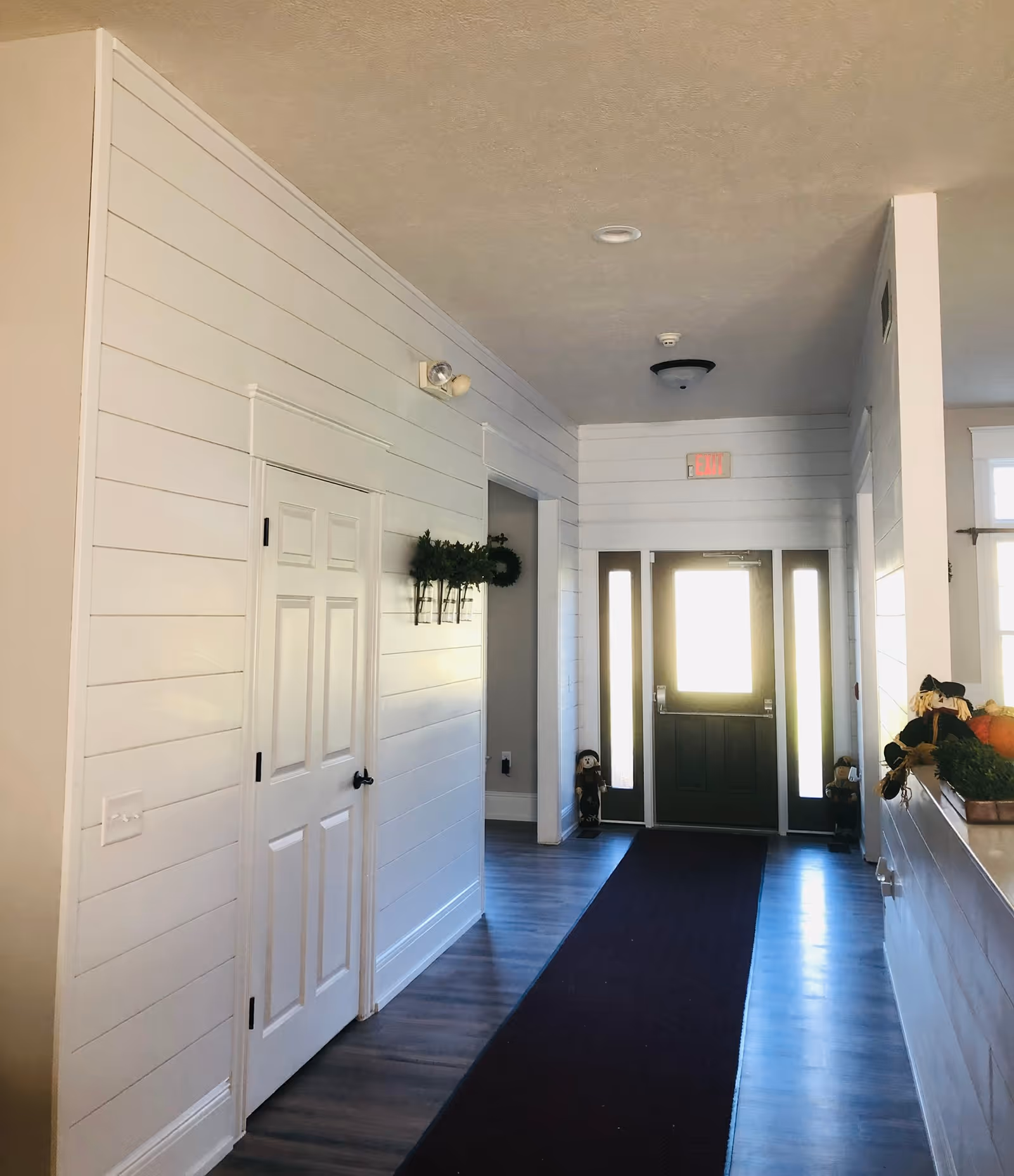 Sunlit entry hallway with white paneled walls, a dark front door at the end, a long dark runner on wood floors and wall decorations.