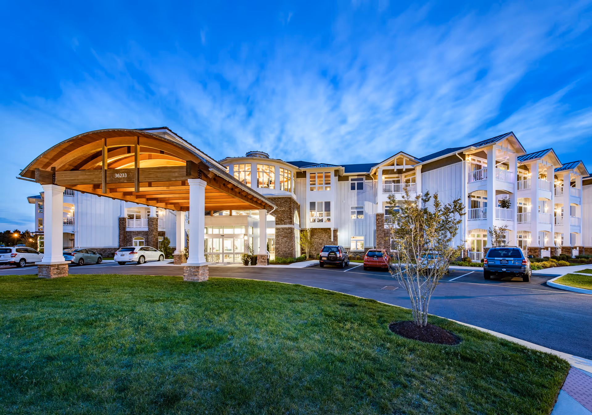 Exterior view of The Lodge at Truitt Homestead building at dusk, featuring a large covered entrance with wooden beams, multiple floors with balconies, parked cars, and a well-maintained lawn and small tree in the foreground under a partly cloudy blue sky.