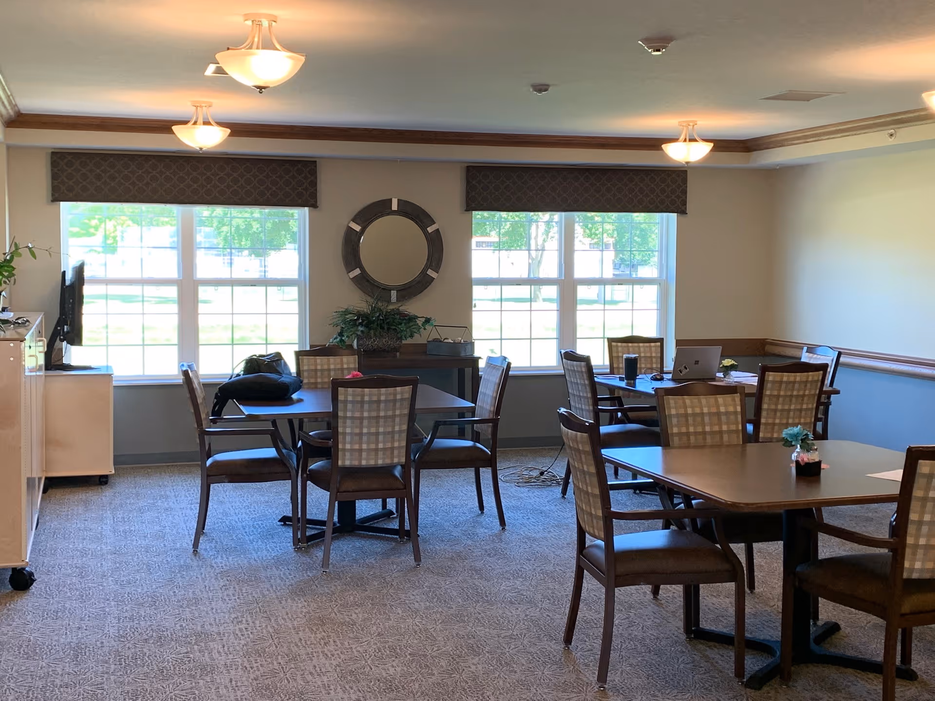 A bright room with multiple tables and chairs arranged for seating. The room has large windows with valances, a round decorative mirror on the wall, and ceiling lights. There are some plants and personal items on the tables and a TV on a cabinet near the window.