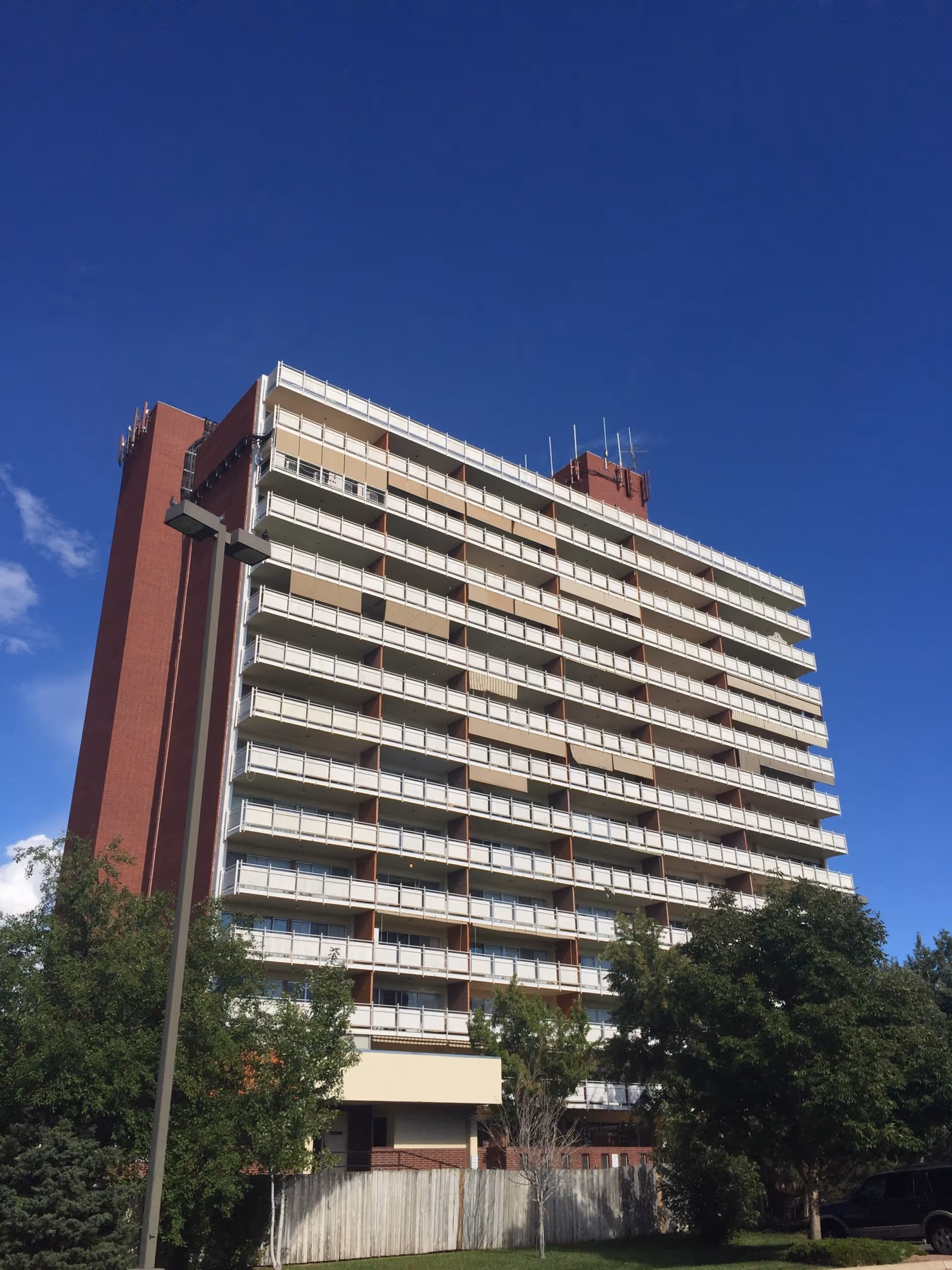 Tall multi-story residential building with balconies on each floor under a clear blue sky, surrounded by trees and a wooden fence in the foreground.