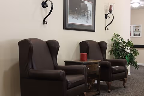 Two dark brown leather wingback chairs with a small wooden side table between them, holding a red candle. Above the chairs is a framed black and white picture of a wheelchair. Two wall-mounted candle holders flank the picture. A green potted plant is visible to the right, and the setting appears to be a quiet indoor hallway or common area.