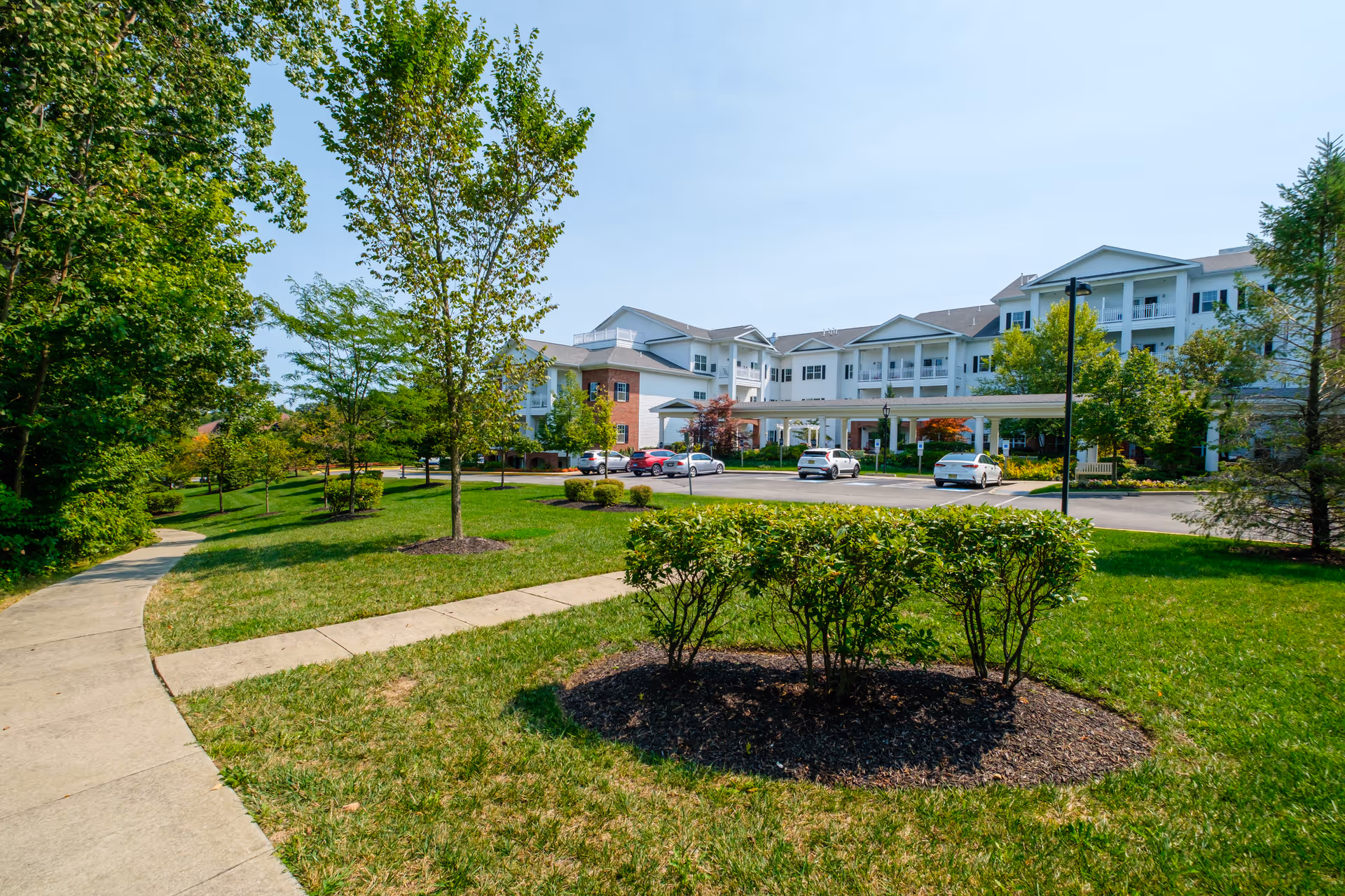 Exterior view of Brightview Randolph senior living facility showing a large white multi-story building with balconies, surrounded by green lawns, trees, and shrubs under a clear blue sky. A paved walkway curves through the landscaped grounds, and several cars are parked near the building entrance.