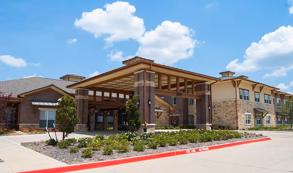 Exterior view of Cedar Bluff Assisted Living & Memory Care building under a blue sky with scattered clouds. The building features a covered entrance with brick pillars, stone and brick facade, and landscaped greenery in front.